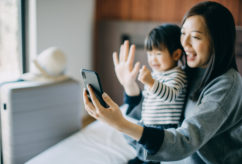 Young Asian mother and cute little daughter having video call on smartphone with family in hotel room while on vacation and smiling joyfully