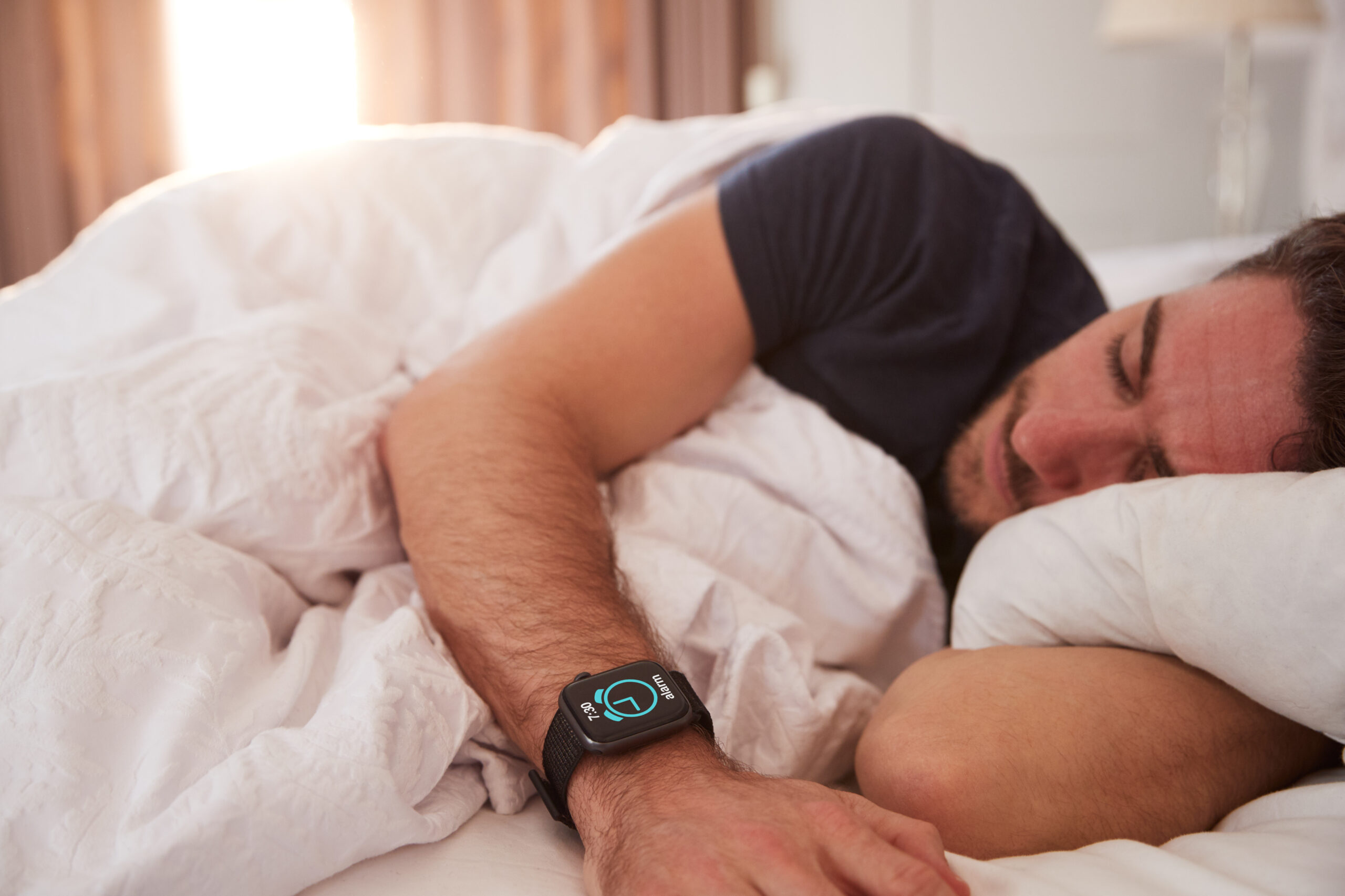 Man Asleep In Bed Wearing Smart Watch As Sun Breaks Through Curtains